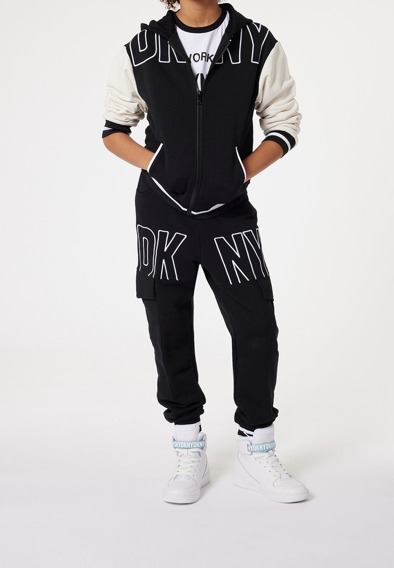 Black and cream zip-up hoodie with large "NY" print, paired with black cargo pants featuring "NY" graphics. White high-top sneakers with accents.