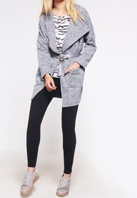 Grå stickad kappa med drapering fram, bälte i midjan, två fickor, lagd över en randig topp, tillsammans med svarta leggings och grå skor.