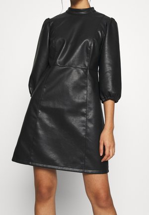 Woman wearing black faux leather knee-length dress with puffed three-quarter sleeves and a high neckline.