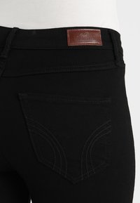 Černé denimové džíny s hnědou koženou visačkou na pásku, s detailním pohledem na design a švy zadní kapsy.