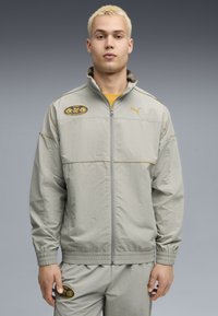 Lys grå sportsjakke med lynlås, elastiske manchetter og en gul accentsnor. Har et sort logo-patch på brystet og matchende bukser.