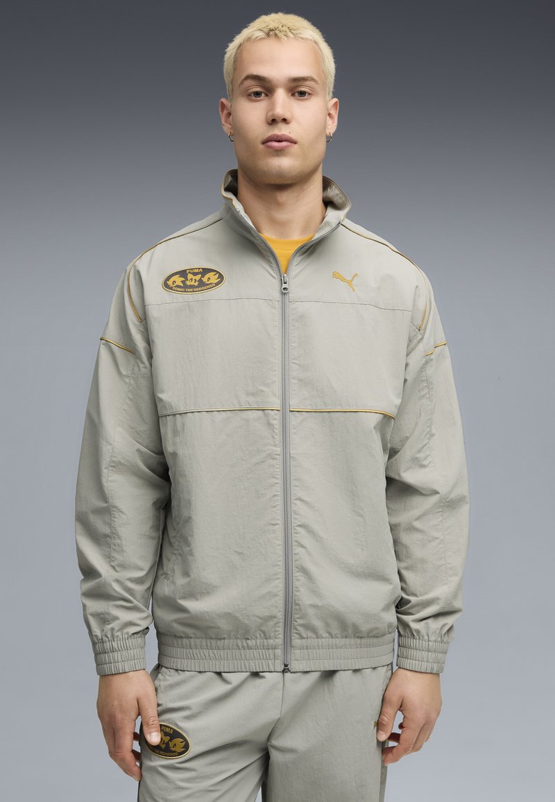 Lys grå sportsjakke med lynlås, elastiske manchetter og en gul accentsnor. Har et sort logo-patch på brystet og matchende bukser.