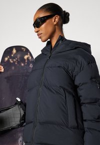 Black puffer jacket with a hood, featuring a front zipper and textured fabric. The model holds a snowboarding board with dark graphics.
