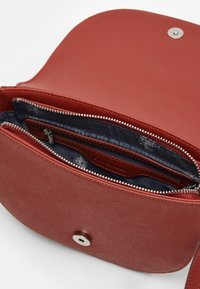 Bolso cruzado de cuero rojo con una textura suave, solapa redondeada, compartimentos con cremallera y cierre con botón plateado. El interior presenta un forro azul marino.