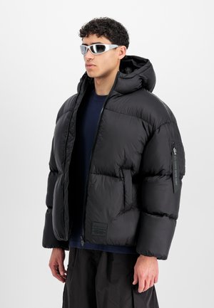 Chaqueta de plumas negra con capucha, cierre de cremallera y varios bolsillos grandes. El material es suave y brillante, con un parche de logo en la parte delantera.