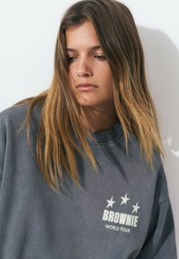 Joven mujer con cabello largo y castaño mira hacia abajo, vestida con una sudadera gris que lleva las palabras "BROWNIE WORLD TOUR" y tres estrellas en el frente.