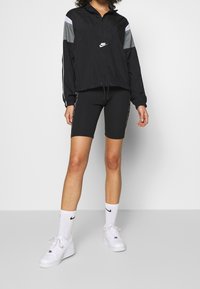 Svart Nike vindjacka med grå accenter, dragkedja vid kragen och dragsko i nederkant, kombinerad med svarta cykelshorts och vita sneakers med logga.