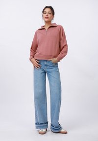Rosa zip-genser med høy krage, vide ermer og ribbestrikket nederkant, kombinert med lyseblå høyhvaste jeans med frynsete mansjetter.