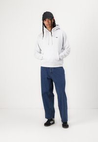Hanorac gri deschis cu glugă, dotat cu un buzunar în față și un logo negru, asortat cu blugi lejeri din denim albastru și adidași negri cu dungi albe.
