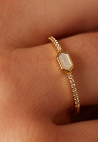Goldring mit einem sechseckigen weißen Stein in der Mitte und einem Band, das mit kleinen klaren Edelsteinen besetzt ist, ruhend auf einer Hand.