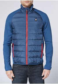 Blaue wattierte Jacke mit eng anliegenden blauen Ärmeln, rotem Reißverschluss und Logo auf der linken Brustseite. Verfügt über Seitentaschen und ein Design mit hohem Kragen.