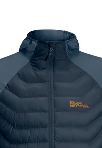 Chaqueta aislante de color azul marino con un alto cuello, patrón acolchado y cierre de cremallera. Presenta paneles en un gris mate contrastante en los hombros. Logotipo de Jack Wolfskin en naranja.