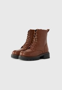 Bottes chevilles en cuir marron avec un design à lacets, bout rond et semelle chunky noire durable. Comprend une languette à l'arrière pour un enfilage facile.