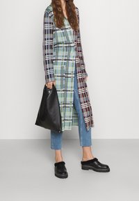 Abrigo de cuadros largo con tonos verdes, azules y morados, usado sobre jeans azul cortos. Bolsa de cuero negra en la mano, combinada con zuecos negros con detalles peludos.