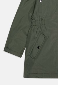 Parka verte avec taille cintrée, accents à boutons-pression et poche latérale. Fabriquée à partir d'un tissu texturé et résistant aux intempéries.