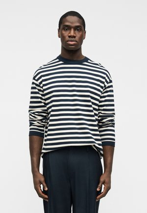 Vīrietis ar tumšu ādu, valkājot garu piedurkņu kreklu ar tumši zilu un baltu horizontālu svītru rakstu un tumšas bikses, stāvošs pret vienkāršu gaišu fonu.