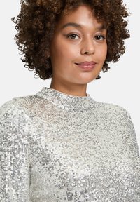 Top z cekinami o wysokim dekolcie, w metalicznym srebrnym kolorze, z teksturowaną powierzchnią i migoczącymi akcentami, które atrakcyjnie odbijają światło.