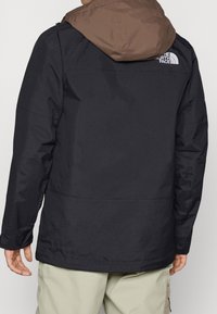 Chaqueta negra con capucha marrón, hecha de un material suave y resistente a la intemperie. Presenta un dobladillo ajustable y un logo en la parte posterior.