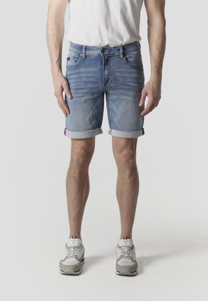 Hombre vestido con pantalones cortos de mezclilla azul claro remangados, zapatillas blancas y camiseta blanca, de pie frente a un fondo liso.