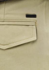 Gros plan d’un tissu beige avec une poche à rabat et une étiquette métallique rectangulaire noire portant la mention « JACK & JONES » près de la poche.