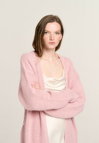 Rosa gestrickte Strickjacke mit langen Ärmeln; weiche, flauschige Textur; lockerer Schnitt; kombiniert mit einem leichten Satin-Top darunter.