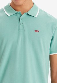 Gros plan sur une personne portant un polo Levi's vert clair avec des bordures blanches au col et aux manches, trois boutons partiellement fermés.