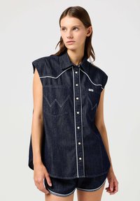 Mørk denimskjorte med spiss krage, korte ermer, to frontlommer og hvite sømdetaljer. Har knapplukking foran.