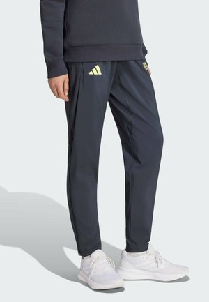 Persona che indossa pantaloni della tuta scuri con logo Adidas giallo, una felpa scura e scarpe da ginnastica bianche, in piedi su uno sfondo chiaro e uniforme.