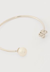 Ανοιχτό ασημένιο βραχιόλι με μεγάλο μαργαριτάρι σε μία άκρη και διακόσμηση από τριφυλλάκι στην άλλη άκρη, πάνω σε λευκή επιφάνεια.