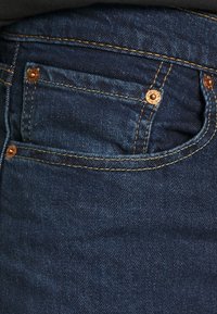 Dark blue denim jeans featuring a small front pocket, visible golden stitching and hardware, with a smooth texture.
