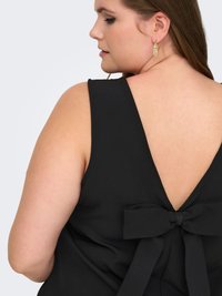 Donna con canotta nera senza maniche con profondo scollo a V sul retro e grande fiocco di tessuto sulla parte bassa della schiena, che mostra il profilo laterale e un orecchino dorato pendente.