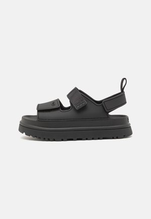 Sandales noires avec une semelle en caoutchouc, des sangles Velcro ajustables et un support au talon arrière, conçues pour le confort et la facilité d'utilisation.