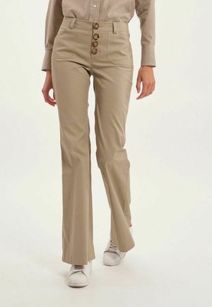 Pantalons évasés beiges avec une taille haute, ornés de quatre grands boutons ronds et de poches latérales. Portés avec des baskets blanches.