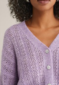 Gaiši lavandas krāsas adīta kardigāna ar V-veida kakla izgriezumu, ar mežģīņu rakstu, lielām pogām un mīkstu, strukturētu apdari.