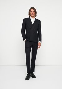 Blazer oscuro, entallado y de corte cruzado con una textura sutil, combinado con pantalones ajustados y mocasines negros, sobre un fondo liso.