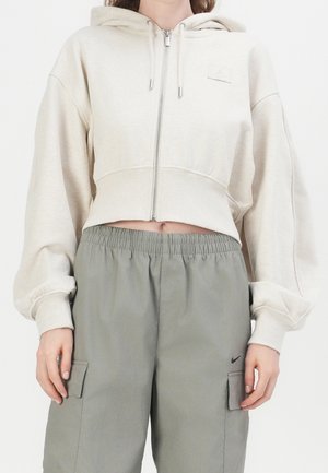 Persona che indossa una felpa beige corta con zip e coulisse, e pantaloni cargo grigi larghi con cinturino elastico in vita e tasche laterali.