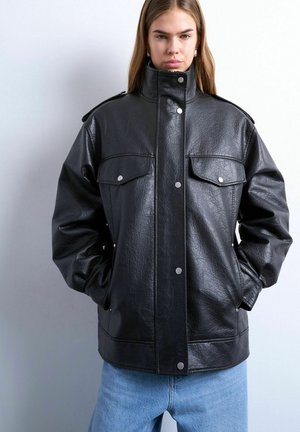 Jonge vrouw met stijl haar draagt een oversized zwarte leren jas met drukknopen en hoge kraag, gecombineerd met lichtblauwe spijkerbroek.