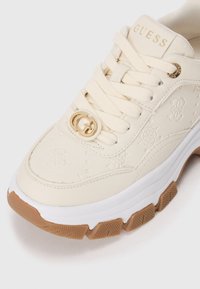 Sneaker di colore crema con motivi in rilievo, suola intermedia bianca, suola in gomma color legno e dettaglio del logo in tonalità oro sui lacci.