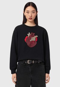 Γυναίκα που φοράει μαύρο φούτερ με λεπτομερή κόκκινη και μπεζ ανατομική καρδιά, συνδυασμένο με μαύρο τζιν και μαύρη ζώνη.