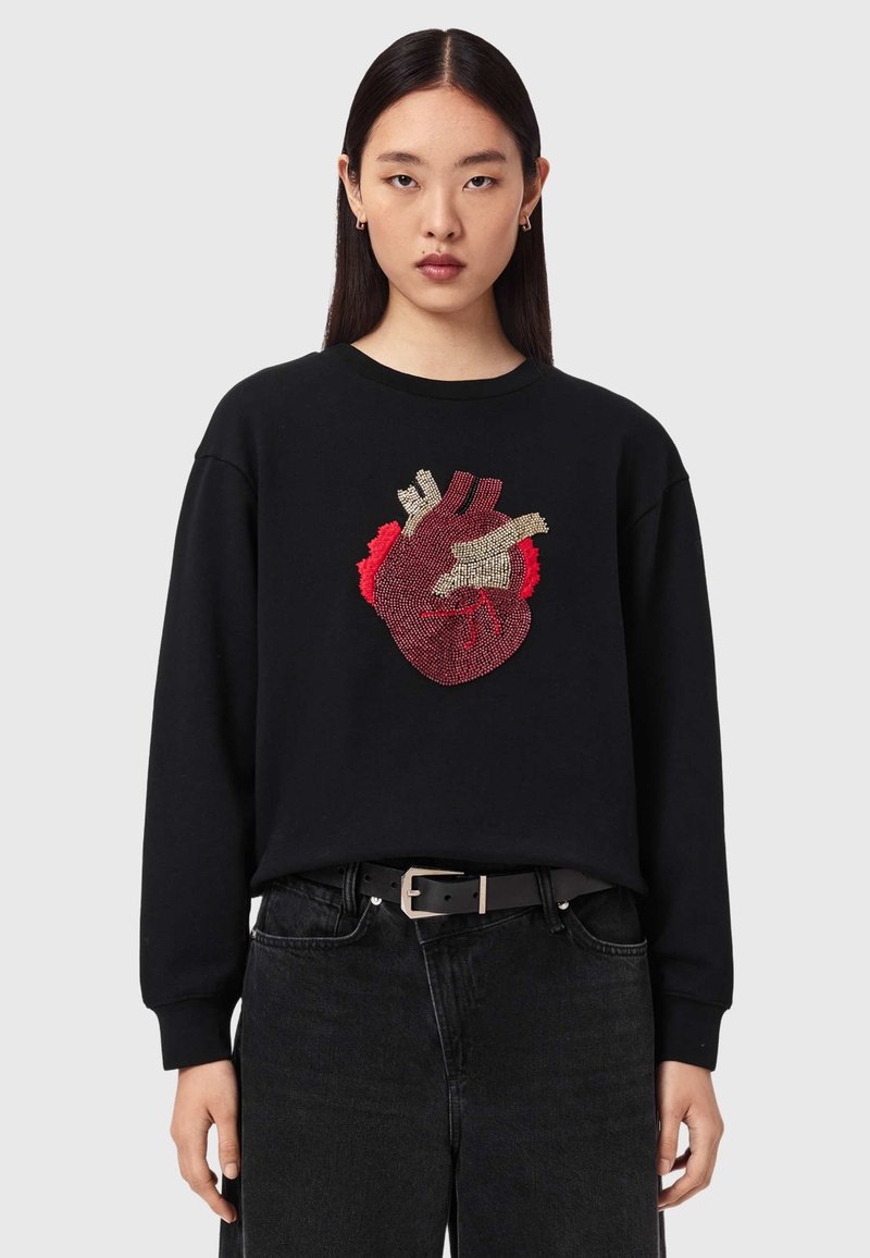 Γυναίκα που φοράει μαύρο φούτερ με λεπτομερή κόκκινη και μπεζ ανατομική καρδιά, συνδυασμένο με μαύρο τζιν και μαύρη ζώνη.