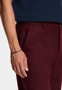 Burgundske hlače iz teksturiranega materiala, opremljene z modrim dekorativnim emblemom, stranskim žepom z zadrgo in zanko za pas z rjavim gumbom.