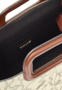 Bolso beige con un patrón sutil, asas de cuero marrón y cremallera dorada. El interior es negro con una etiqueta "TOUS" cosida en su interior.