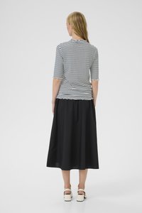 Frau mit langen blonden Haaren, die ein schwarz-weiß gestreiftes Oberteil mit horizontalen Streifen, einen schwarzen Midi-Rock und weiße Sandalen mit klobigen Absätzen trägt und mit dem Rücken zur Kamera steht.