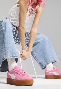 Osoba s růžovými vlasy sedí na bílé drátěné židli pozpátku, má na sobě světlé modré široké džíny a růžové platformové tenisky Puma.