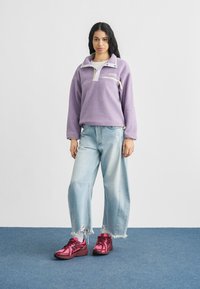 Μωβ φλις πουλόβερ με μισό φερμουάρ, λευκό γιακά και πλαϊνή τσέπη. Συνδυασμένο με ανοιχτό μπλε τζιν παντελόνι με φαρδιά πόδια και ροζ αθλητικά παπούτσια.