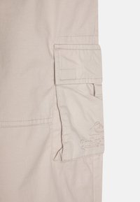 Beige cargo nadrág, oldalzsebbel és fedéllel, varrott részletekkel és sima pamut textúrával.