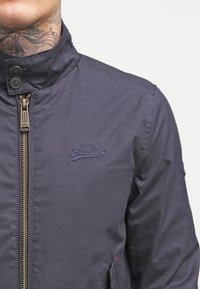 Tmavě modrá bunda s vysokým límcem, zapínáním na zip a logem na hrudi. Má hladkou texturu a zlatě tónovaný zip.