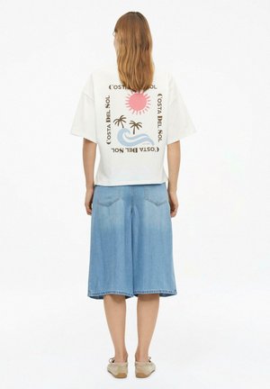 Frau trägt weißes T-Shirt mit pinker Sonne, Palmen, blauen Wellen und dem Schriftzug „Costa del Sol“, kombiniert mit einem knielangen Jeansrock.