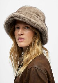 Brauner Kunstfell-Bucket-Hut mit breiter Krempe und weicher Textur, kombiniert mit einer braunen Lederjacke. Dezente Nähte betonen das Design.