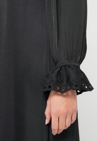 Black dress close-up featuring long, pleated sleeves with gathered cuffs. Cuffs are adorned with circular cutouts, showcasing a textured fabric.